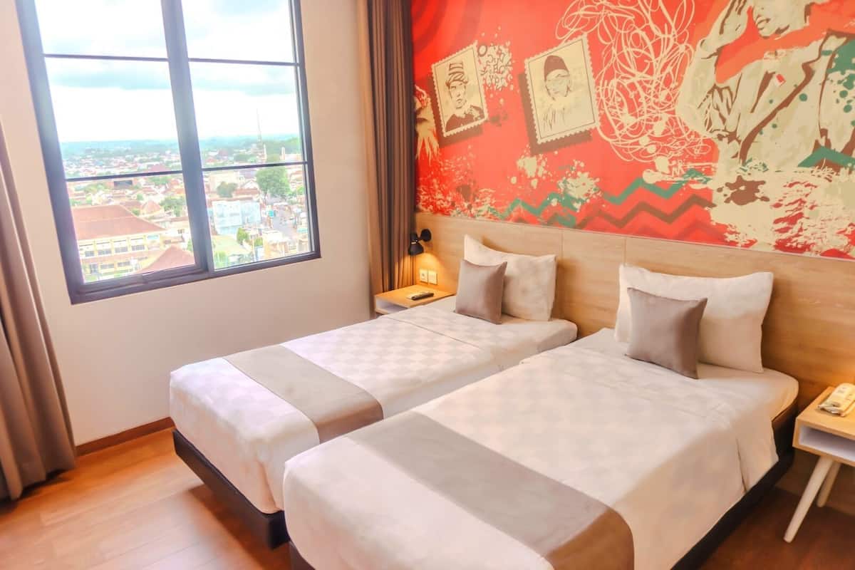 A hotel room with two beds, a large window with a city view, a red wall with artwork, and a nightstand with a lamp.