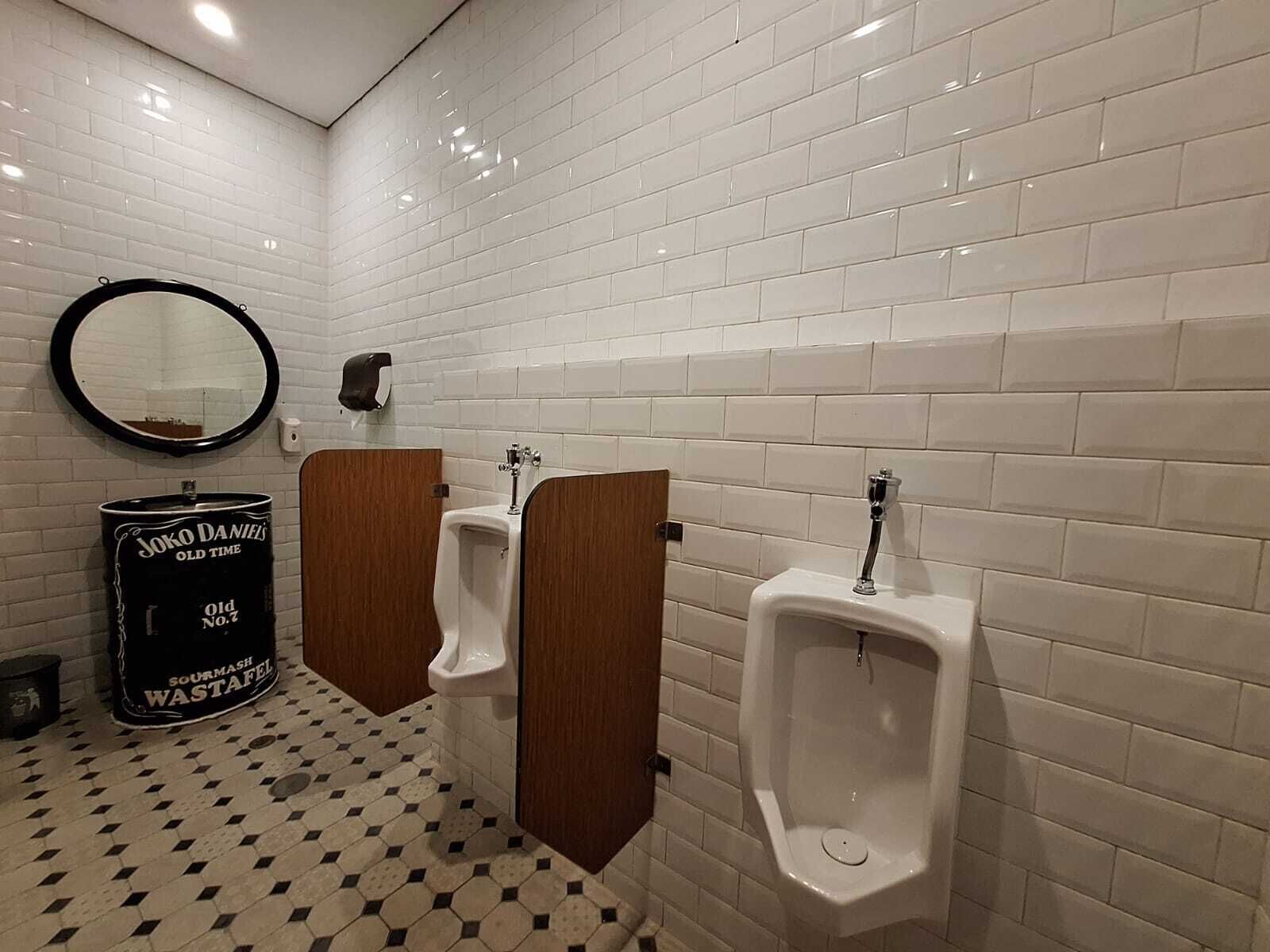 A men's restroom with white tiled walls, a mirror, two urinals, and a black trash can labeled "Joko Daniels Old Time Sour Mash Wastafel".