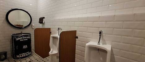 A men's restroom with white tiled walls, a mirror, two urinals, and a black trash can labeled "Joko Daniels Old Time Sour Mash Wastafel".
