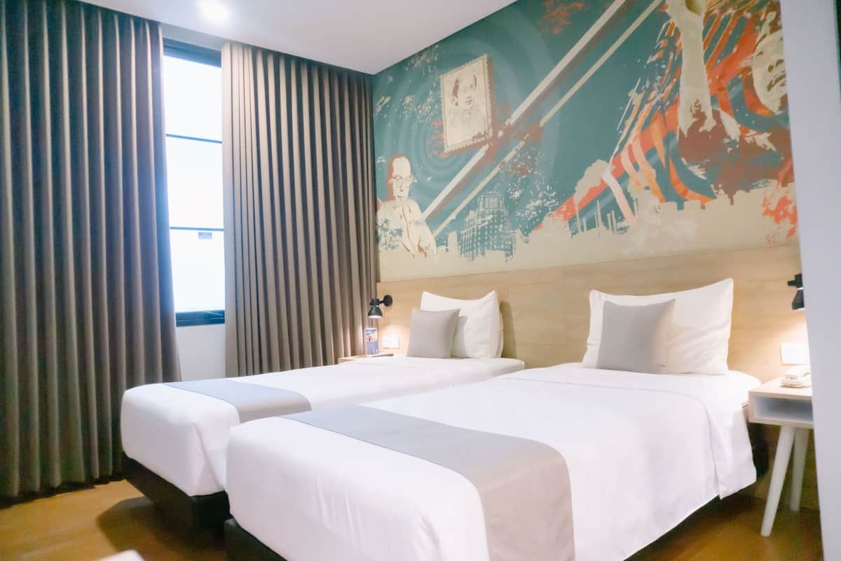 A hotel room with two beds, a large window with curtains, a mural on the wall, and a nightstand with a lamp.