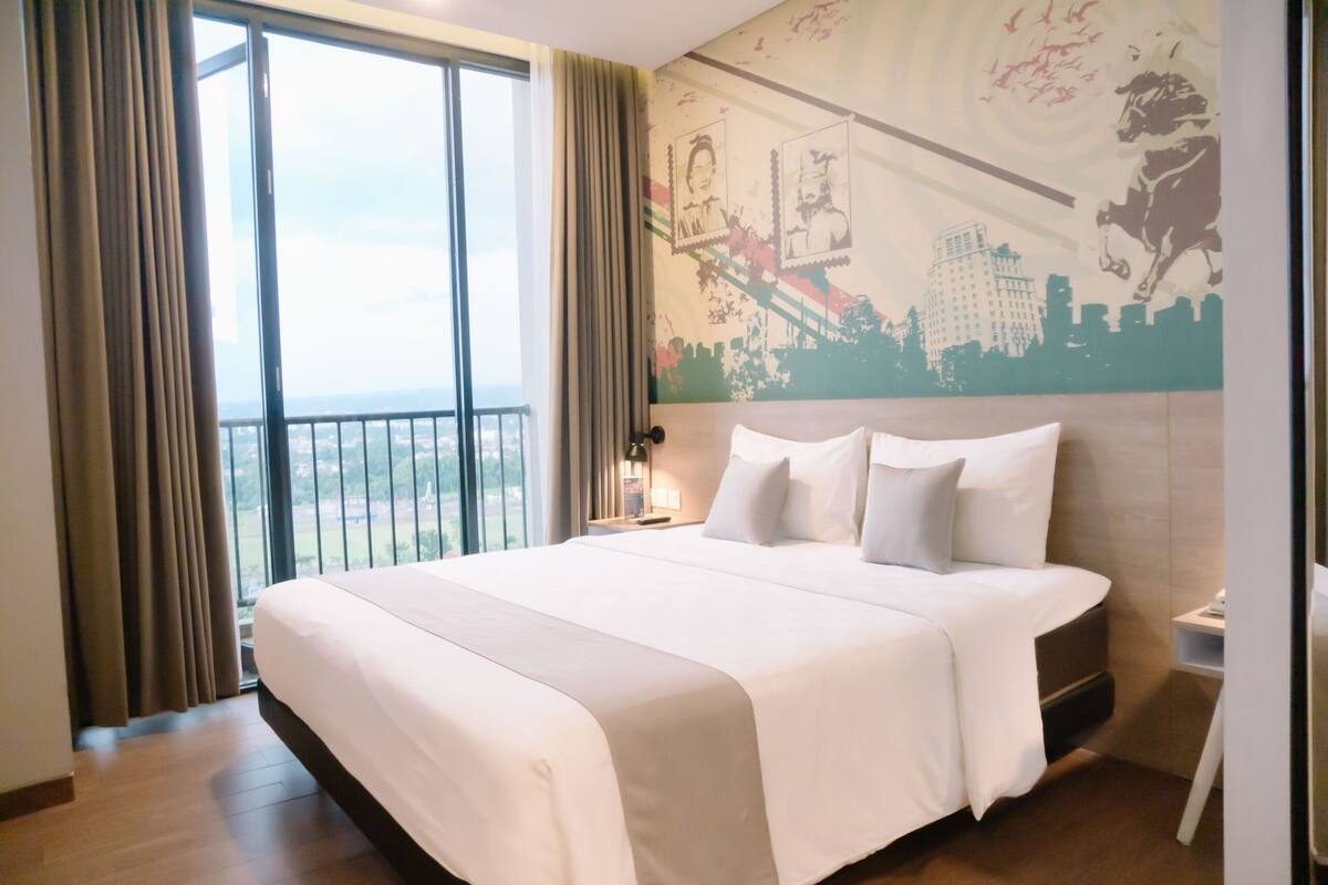 A hotel room with a large bed, a nightstand, a lamp, and a view of the city through a large window.