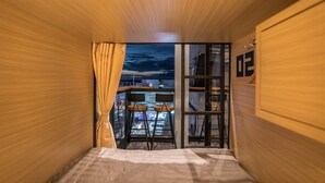 A room with a bed, a window with curtains, a view of the cityscape, and a small seating area with stools. - Sleep Box Patong - Hostel (Patong)