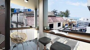 A balcony with a wicker chair, a black stool, and a view of a cityscape with buildings and a clear sky. - Sleep Box Patong - Hostel (Patong)