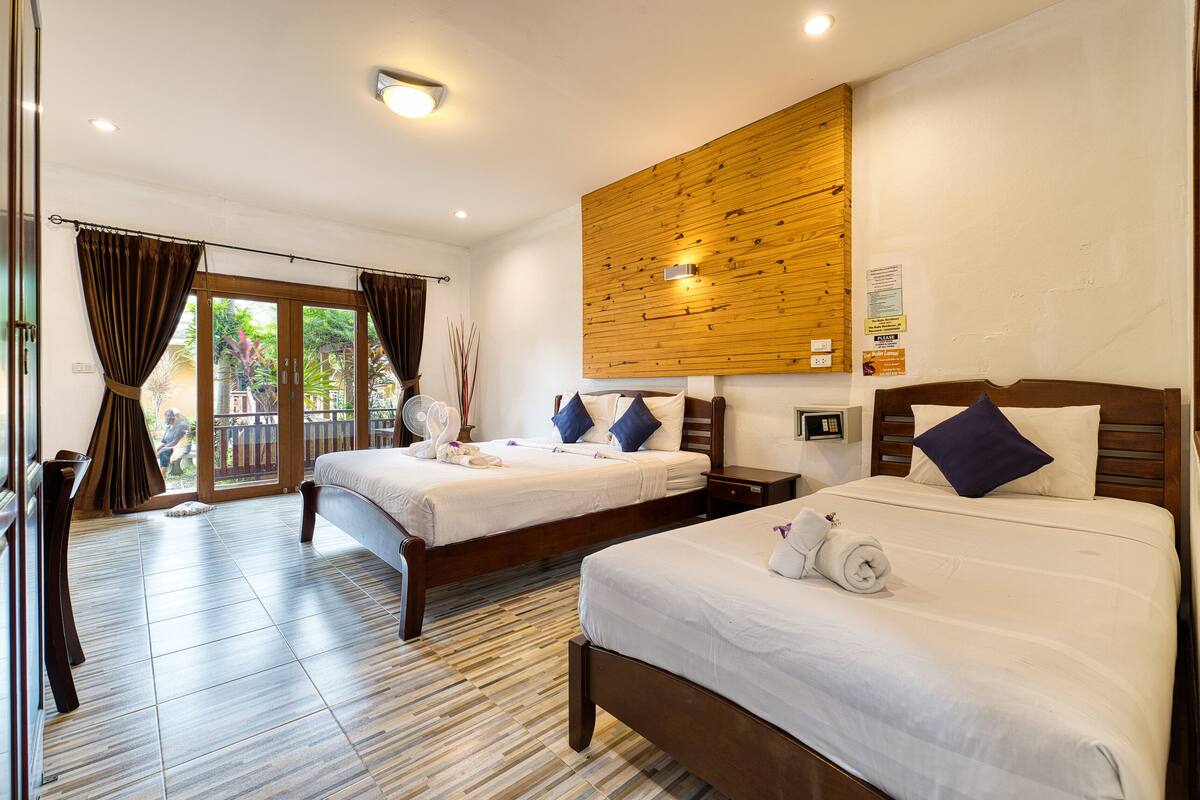 A hotel room with two beds, a wooden wall panel, a balcony with curtains, and a tiled floor.