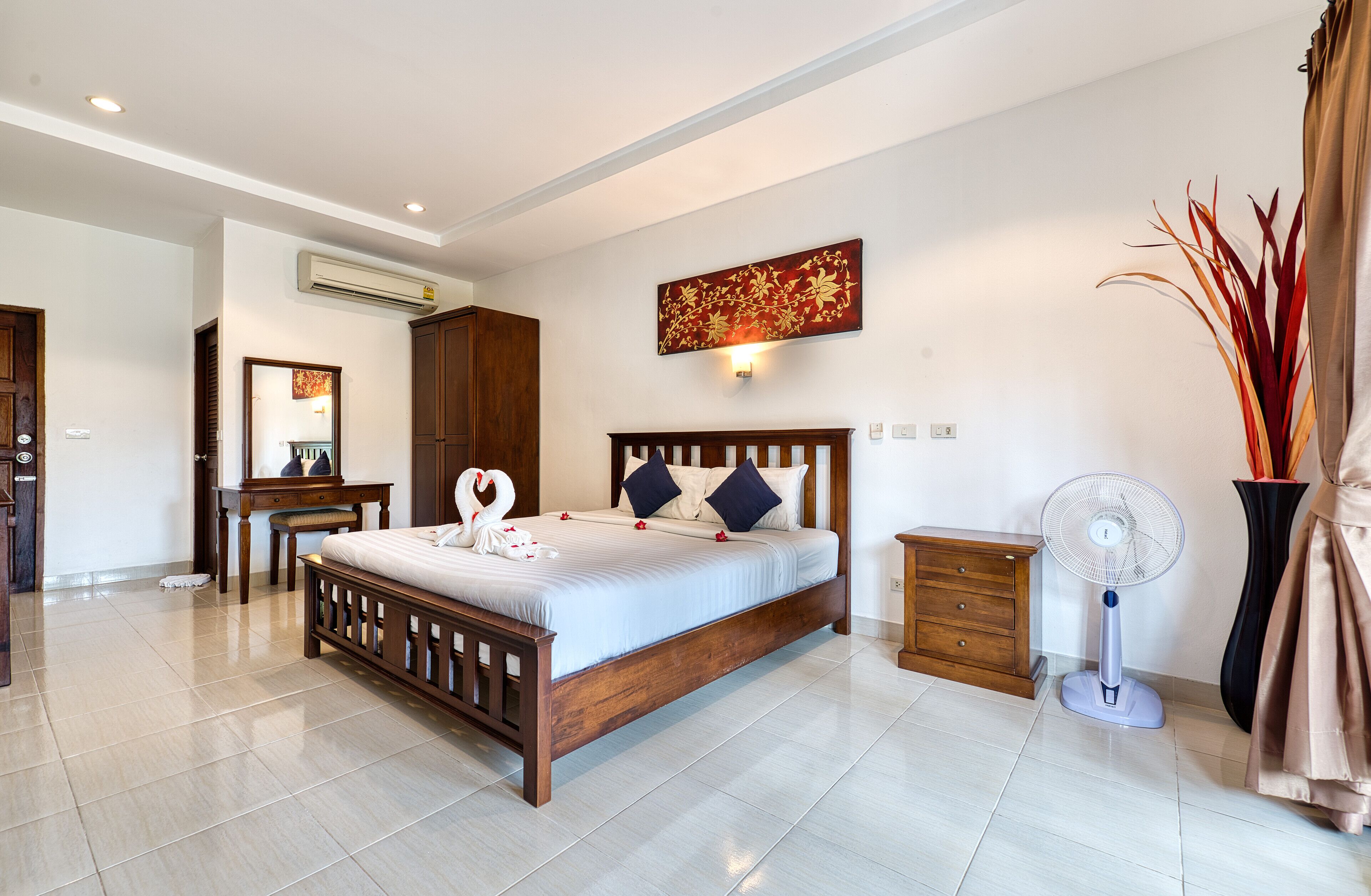 A bedroom with a wooden bed, a nightstand, a wall-mounted air conditioner, a standing fan, and a decorative wall piece.