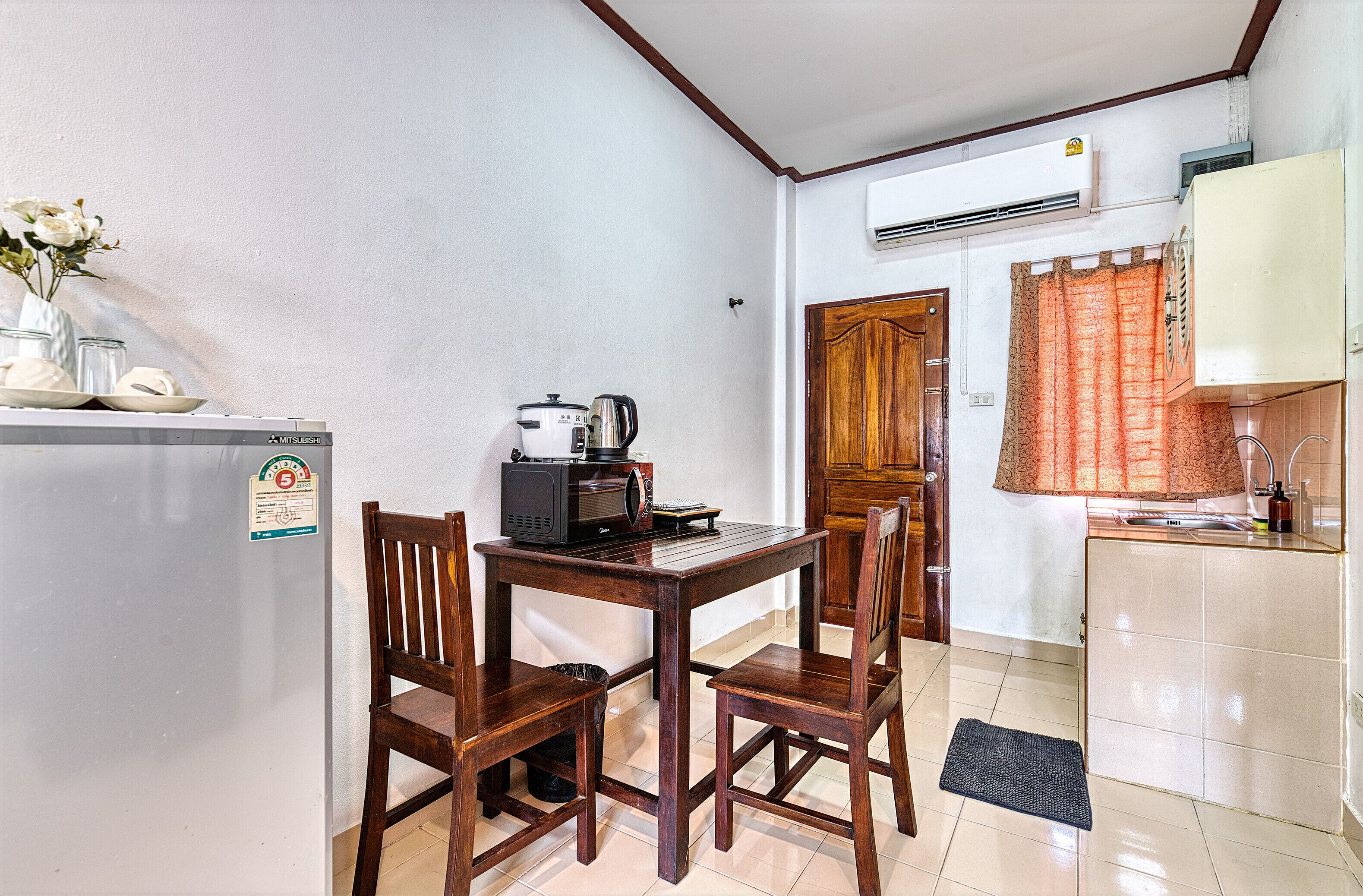 A compact kitchen with a refrigerator, microwave, and stove. There is a wooden dining table with chairs. The kitchen has a tiled floor and a window with curtains.