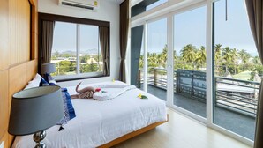 Hotellihuone, jossa on suuri sänky, yöpöytälamppu, näkymä parvekkeelle ja ikkuna, josta avautuu näkymä palmuille.