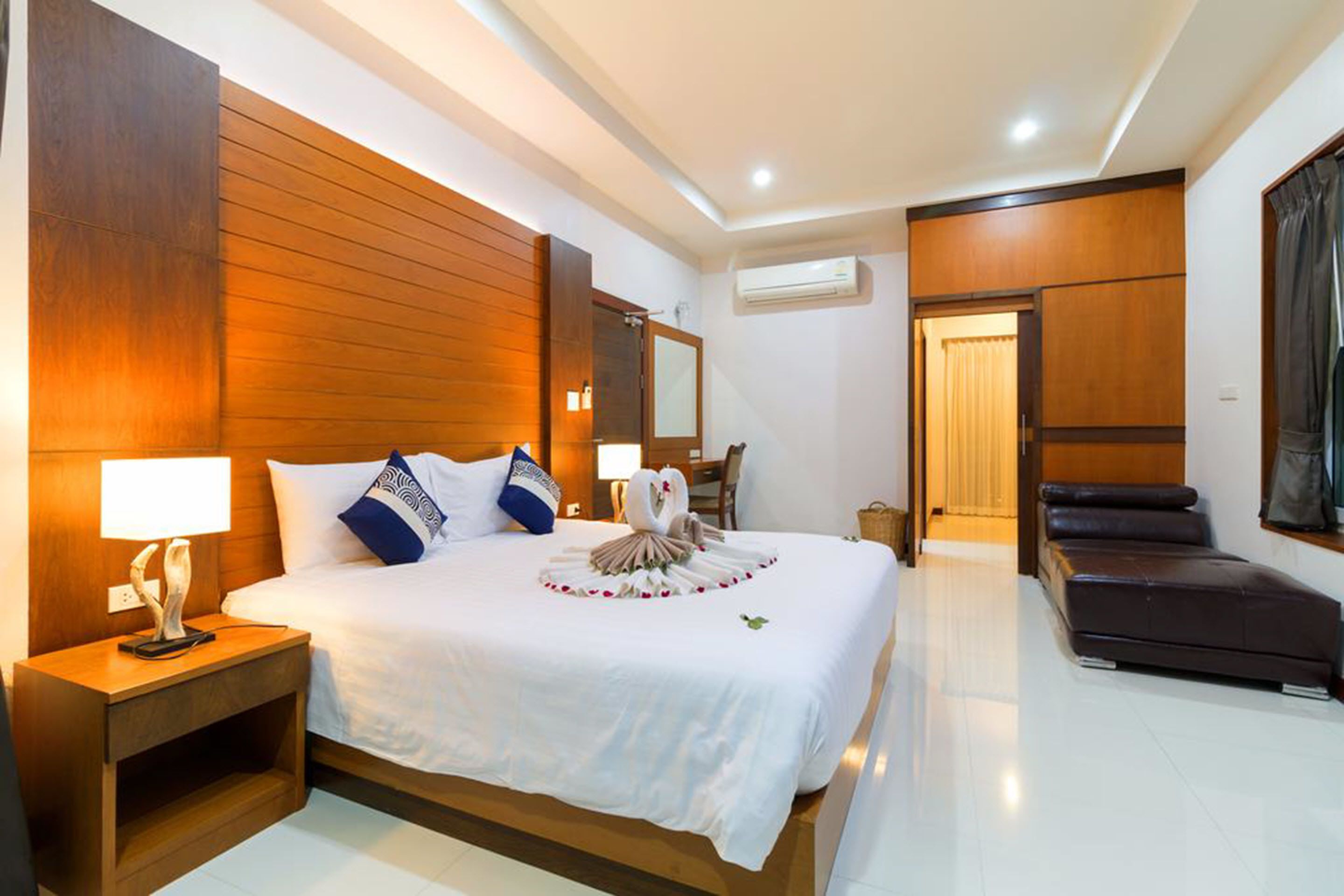 A hotel room with a large bed, wooden headboard, bedside tables, a mirror, and a seating area with a sofa and armchair.
