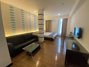 A modern hotel room with a sofa, a glass coffee table, a bed with a blue bedspread, a TV on a stand, and a wall with a geometric pattern.