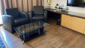 Una moderna camera d'albergo con un tavolino in vetro, due divani in pelle, un armadietto in legno, una TV a schermo piatto e una lampada.