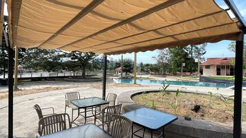 Un espacio patio al aire libre con mesas y sillas bajo un toldo, una piscina al fondo y un jardín ajardinado.