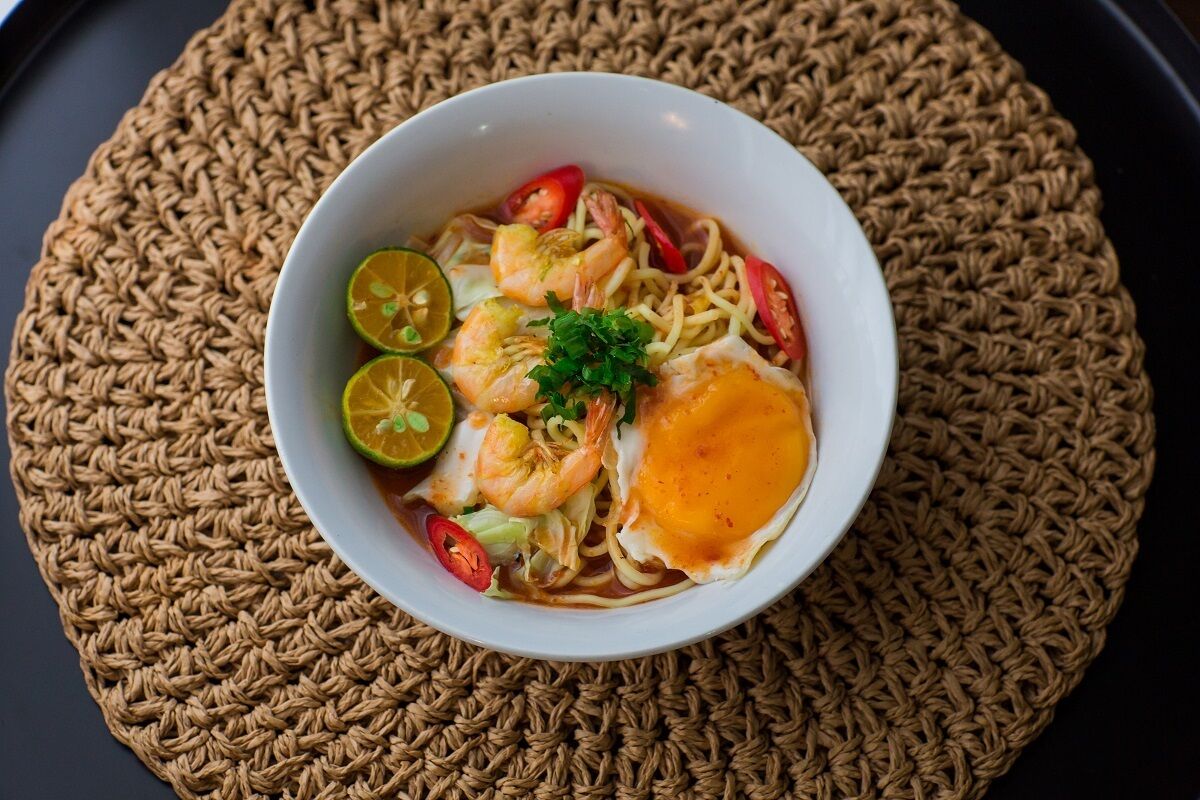A bowl of noodle soup with shrimp, egg, and lime on a woven placemat.