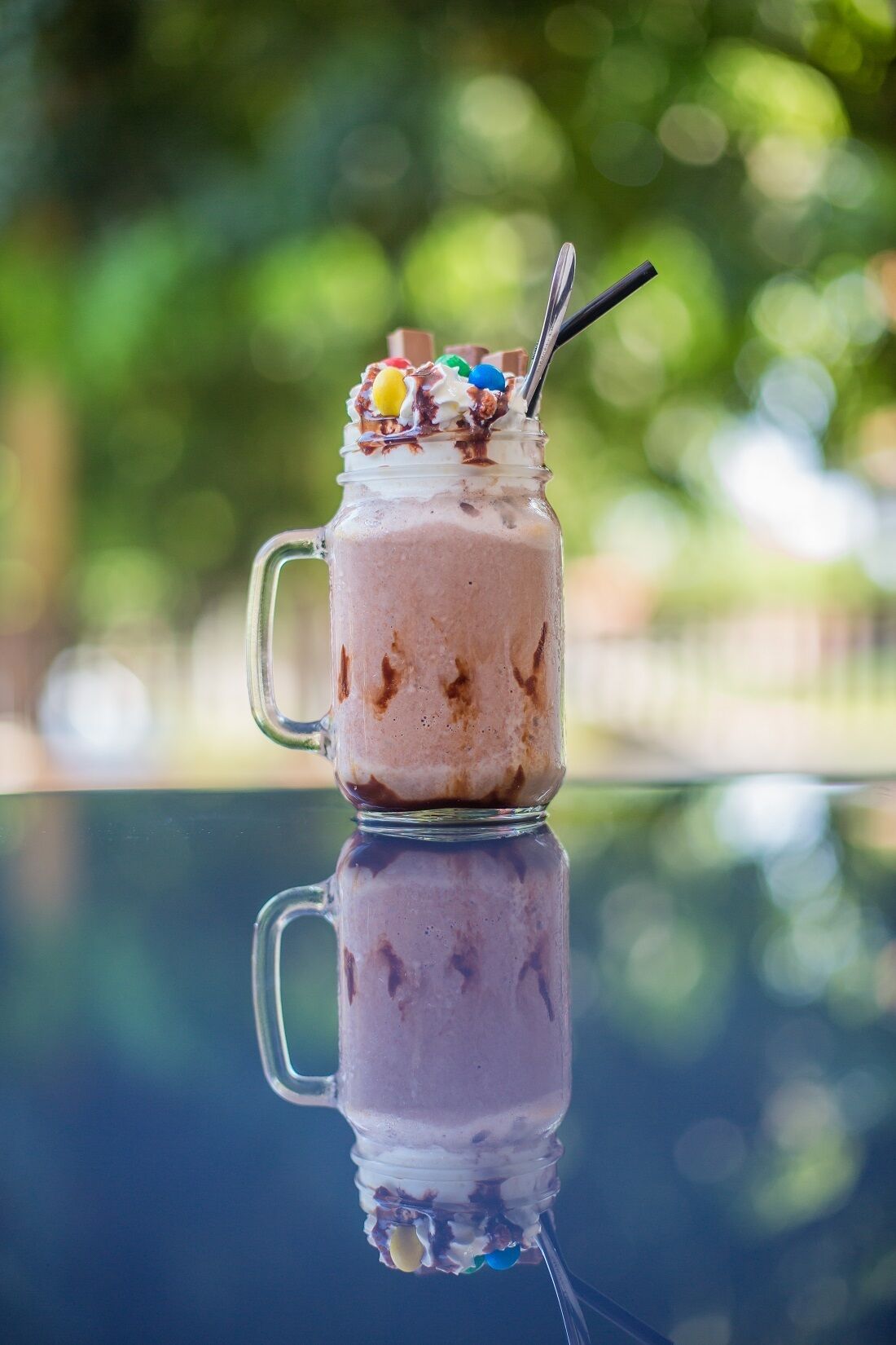 A milkshake in a glass jar with a straw, topped with whipped cream and colorful candies, placed on a reflective surface.