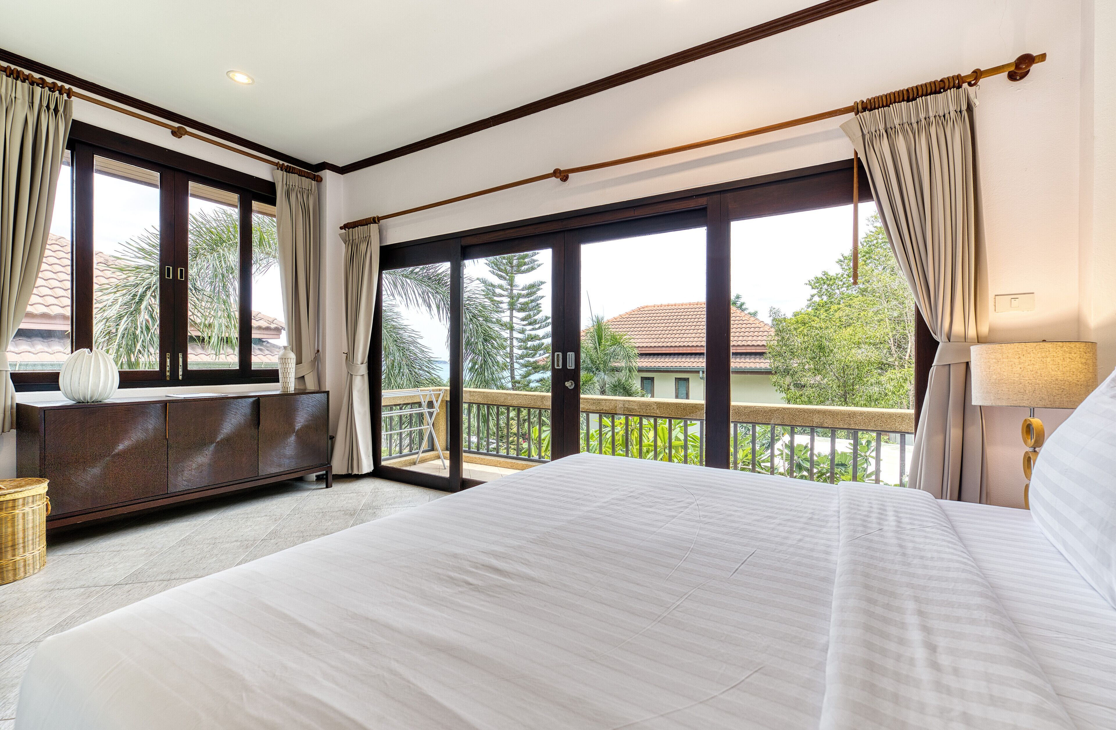 A hotel room with a large bed, a dark wooden cabinet, a wicker basket, and a balcony with a view of palm trees and buildings.