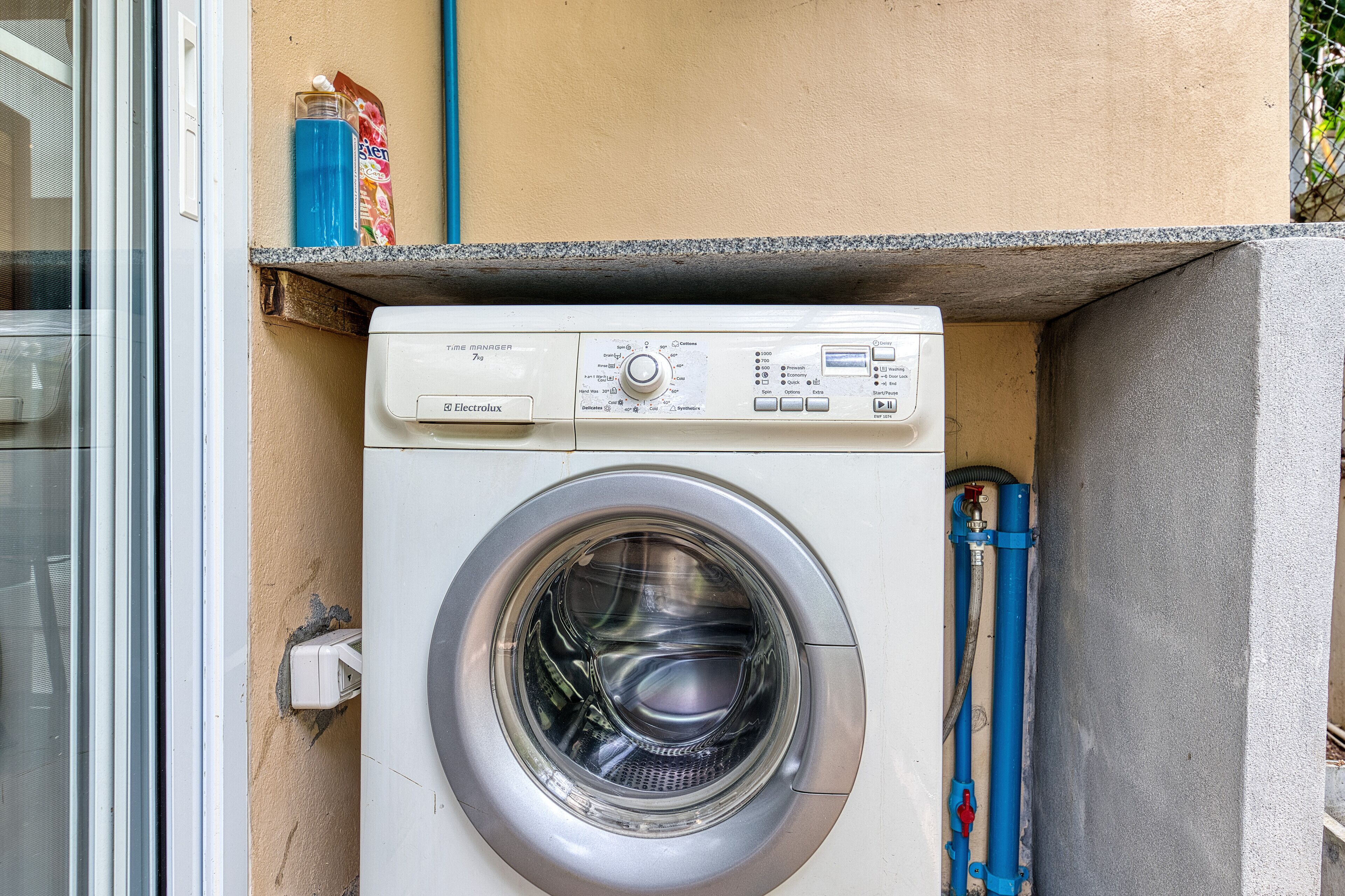 A front-loading washing machine installed in a corner of a building, with a control panel and a hose connected to it.