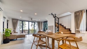 A spacious living area with a wooden dining table, comfortable seating, a staircase, and large windows.