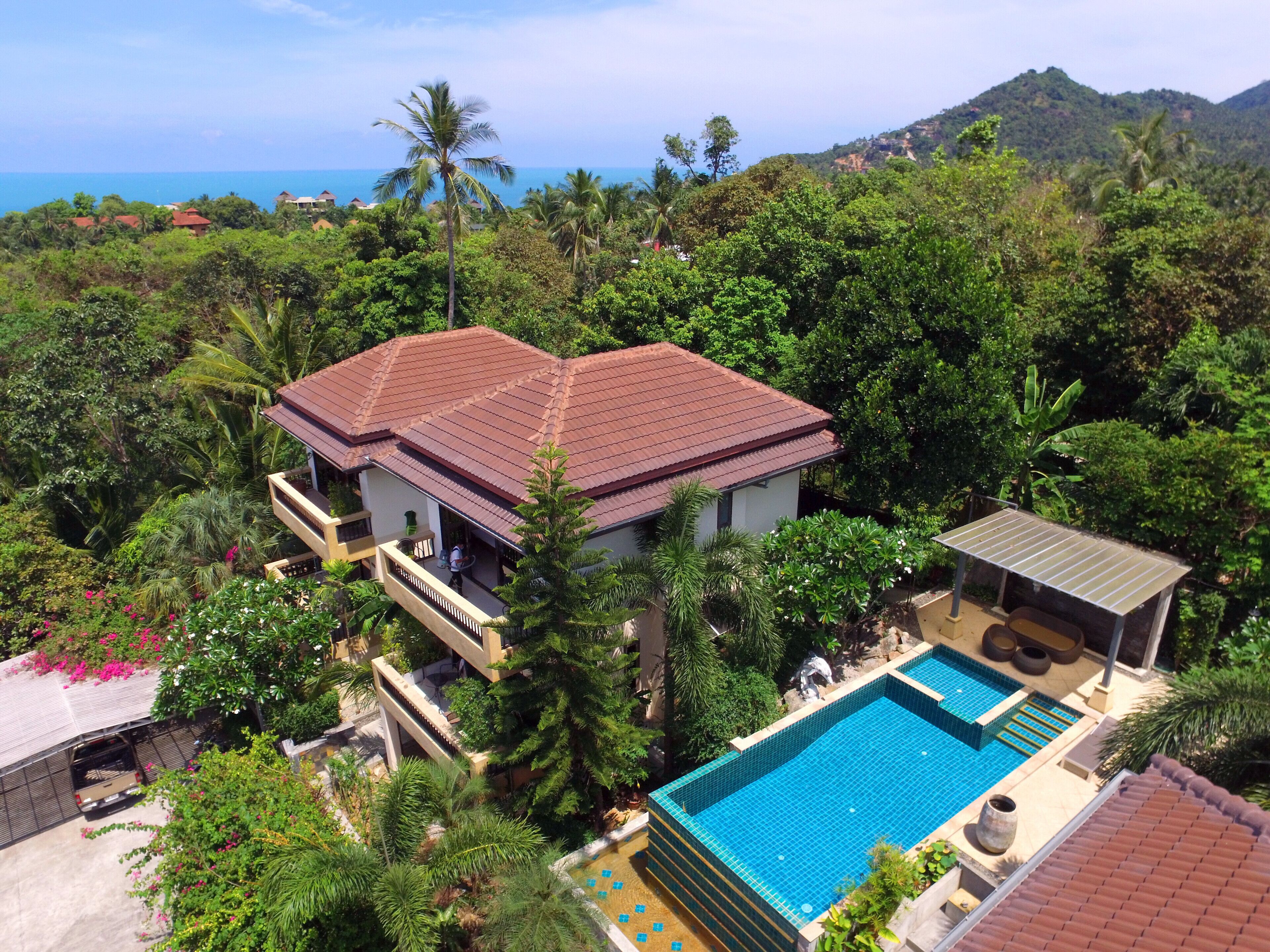 A two-story house with a swimming pool, surrounded by lush greenery and a view of the ocean.
