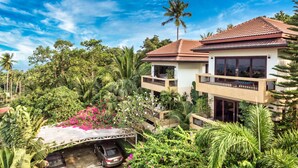 A two-story house with a red-tiled roof, surrounded by lush greenery and a clear blue sky.