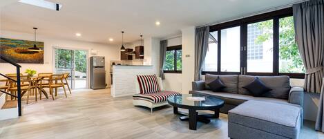 A modern living room with a sofa, ottoman, and coffee table, a dining area with a table and chairs, and a kitchen with a refrigerator and pendant lights.
