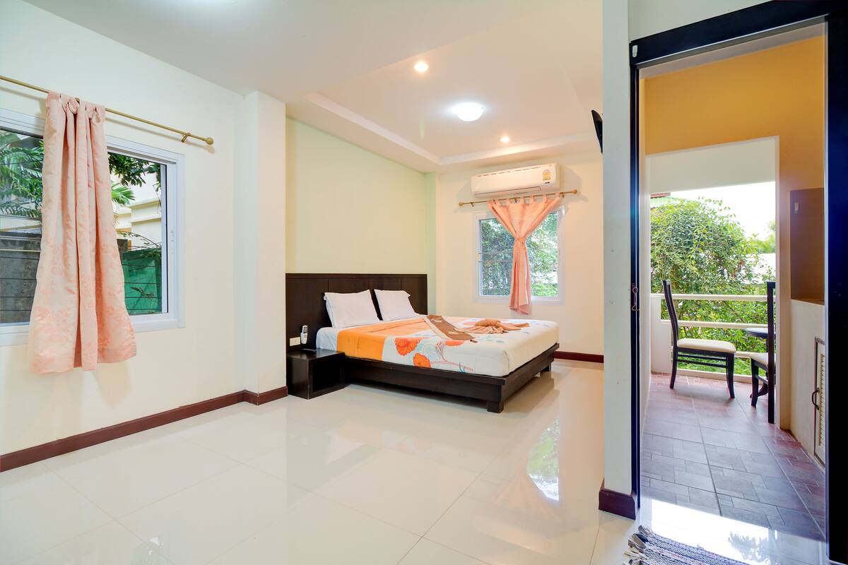A bedroom with a bed, a window with curtains, an air conditioning unit, and a balcony with a chair.
