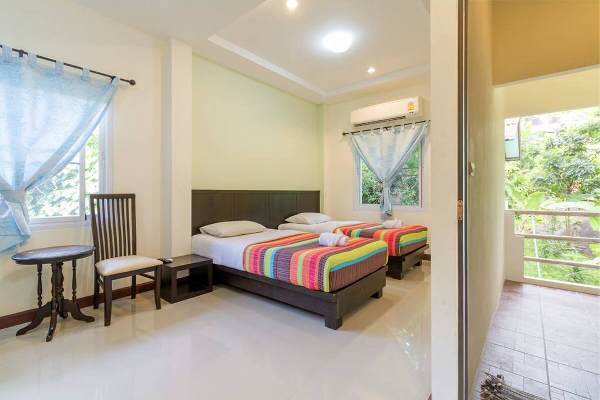A hotel room with two beds, a chair, a small table, and a balcony with a view of greenery.