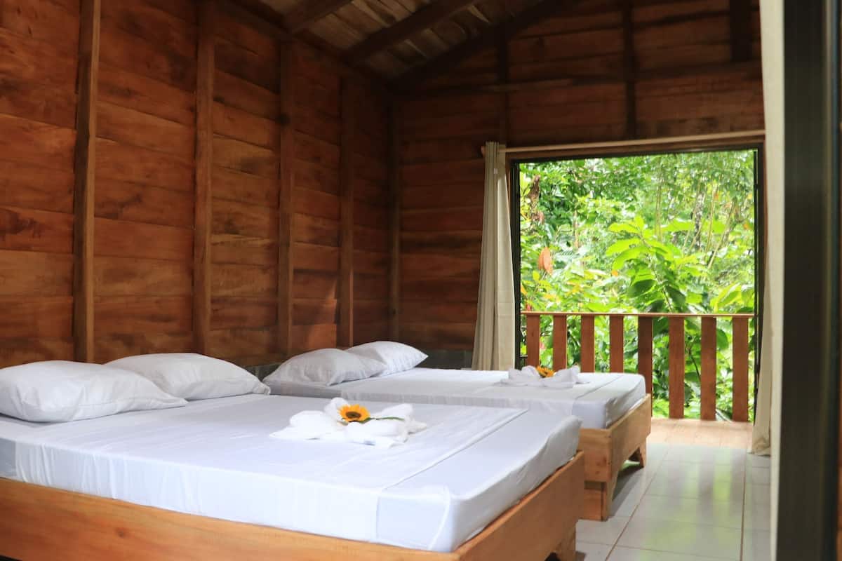 A wooden cabin with two beds, white linens, and a balcony with a view of greenery.
