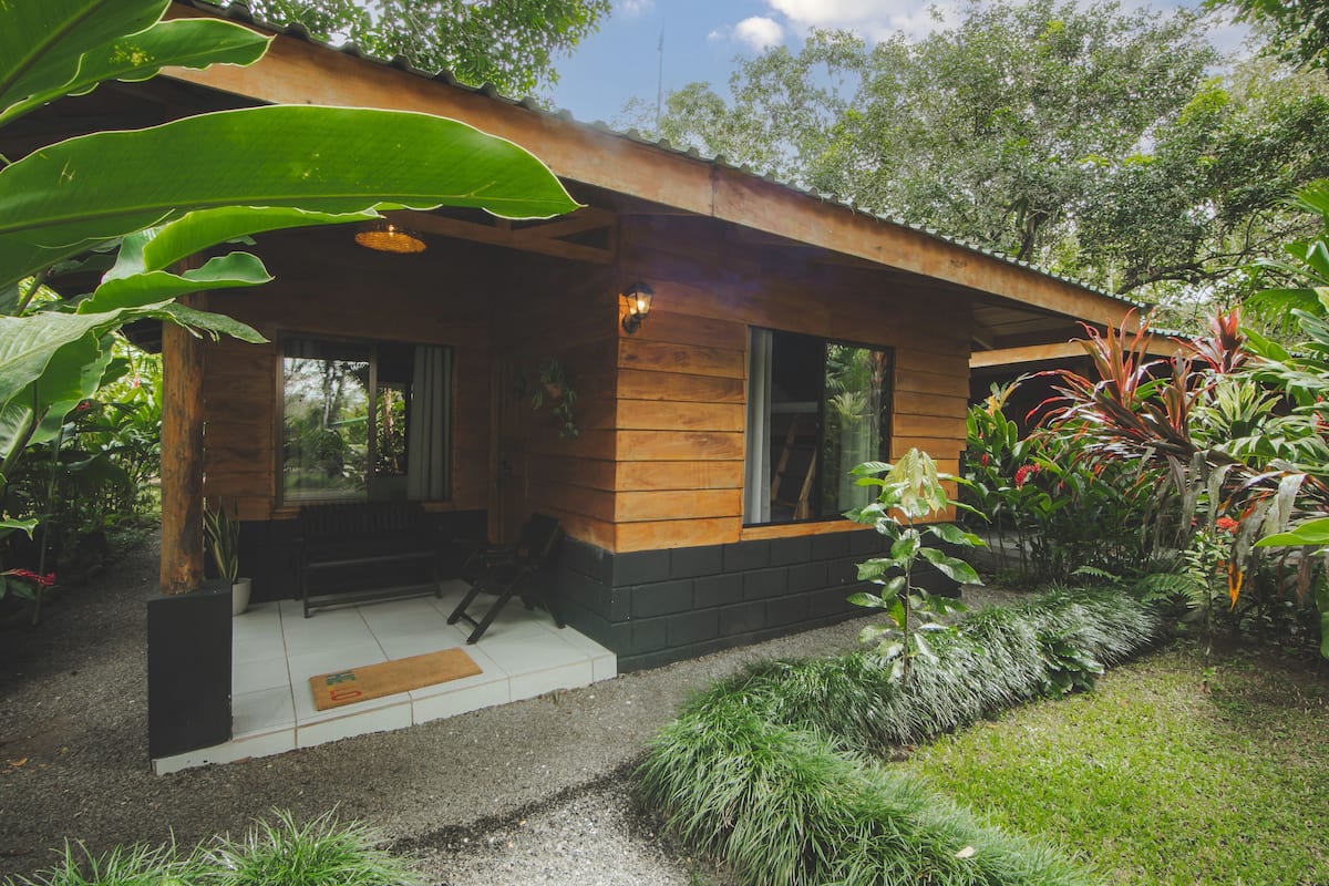 A small wooden cabin with a porch, surrounded by lush greenery.