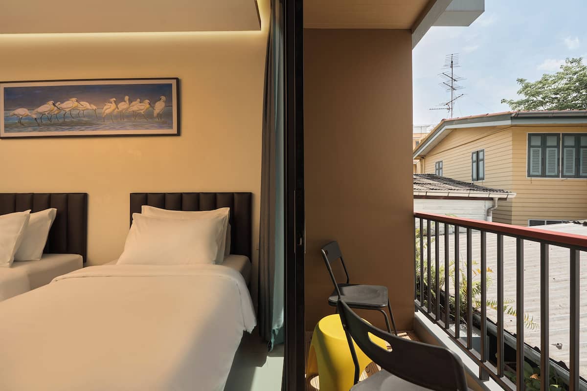 A hotel room with two beds, a balcony with a yellow table and chairs, and a painting of birds on the wall.