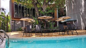 Una piscina con sillas de descanso y sombrillas, rodeada de plantas y un edificio con grandes ventanales.