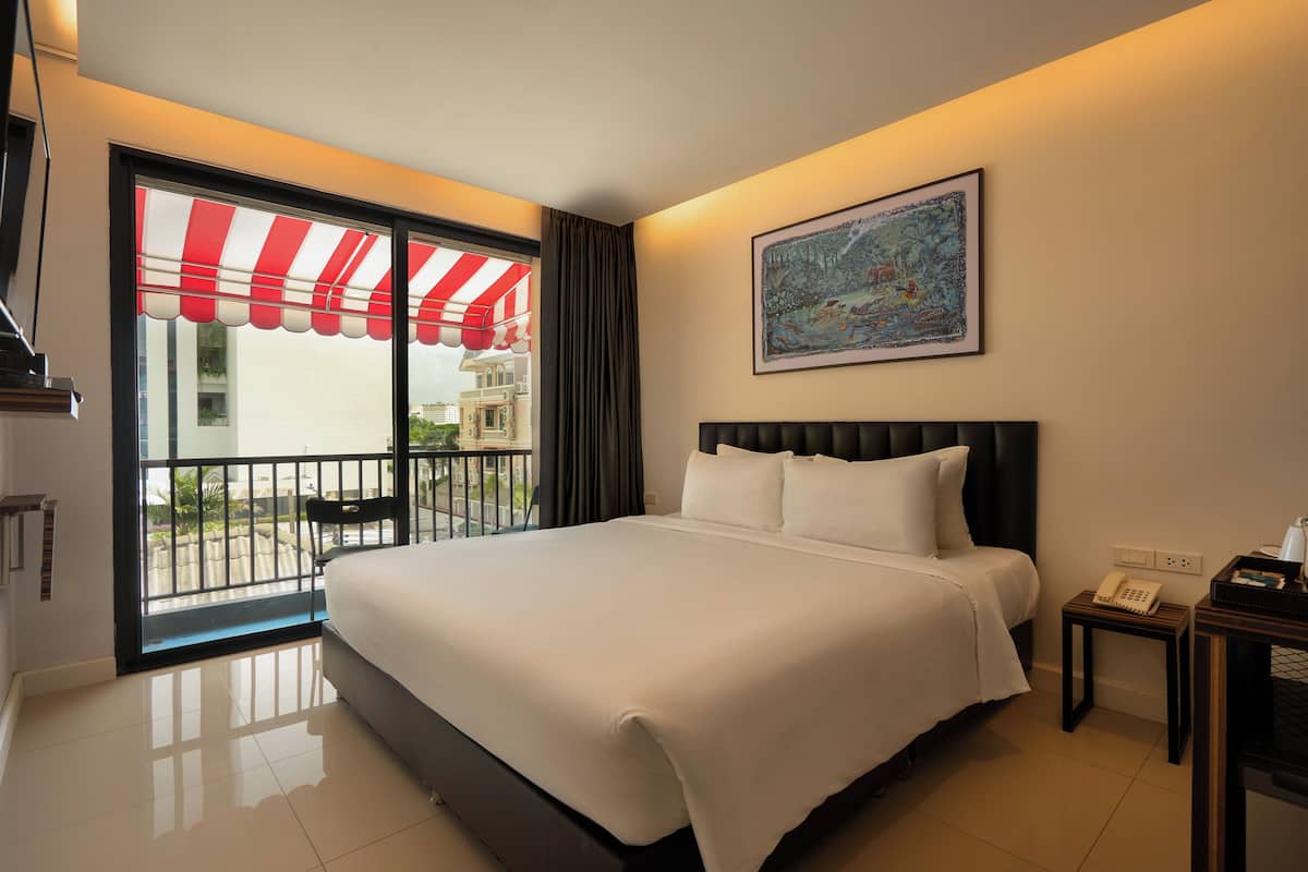 A hotel room with a large bed, a painting on the wall, a balcony with a red and white striped awning, and a view of buildings outside.