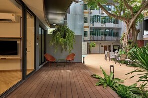 Un área de terraza al aire libre con piso de madera, mobiliario y un gran árbol que ofrece sombra.