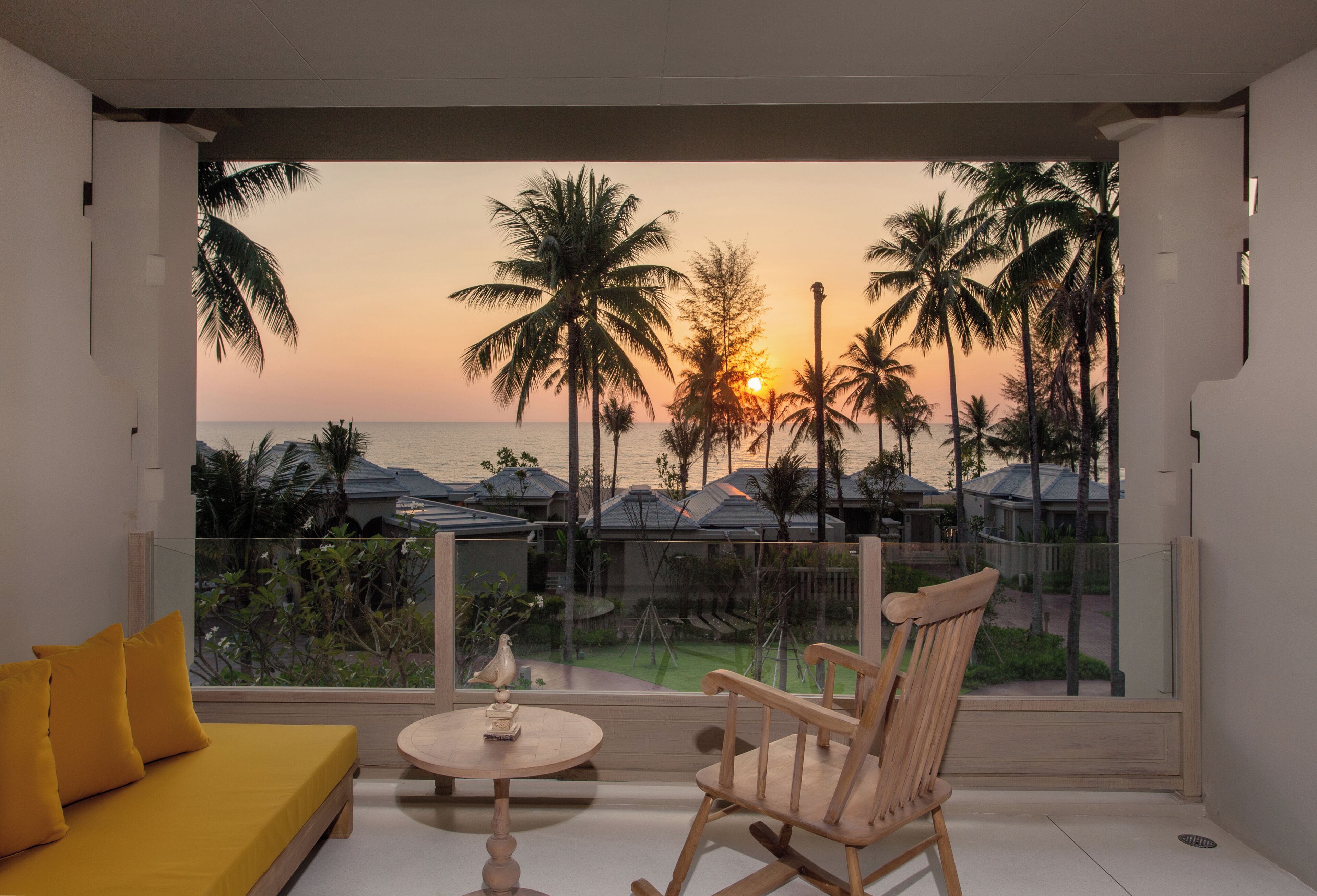 Ein Balkon mit Blick auf Palmen und den Sonnenuntergang, ein gelbes Sofa und ein hölzerner Schaukelstuhl.