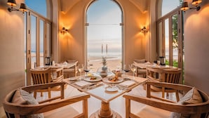 A dining area with tables and chairs, a view of the beach, and a large arched window. - Devasom Khao Lak Beach Resort & Villas (Takua Pa)