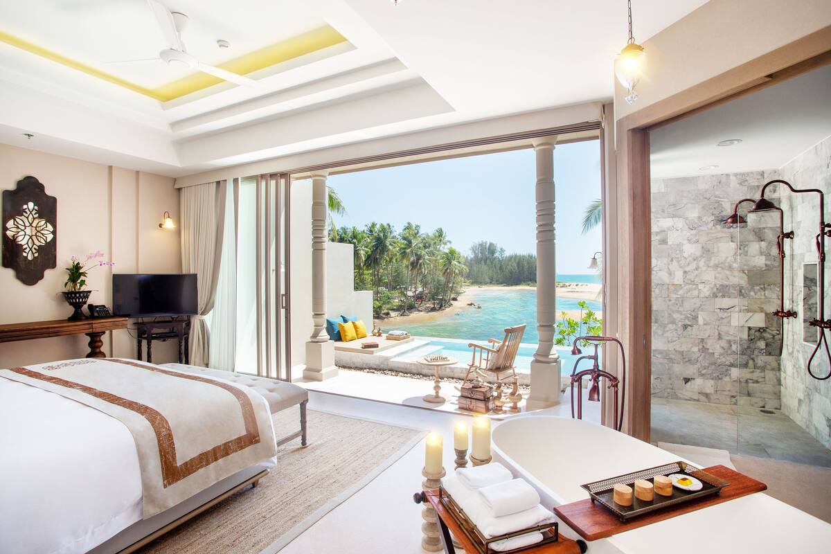 A hotel room with a bed, a TV, a bathroom with a shower, and a view of the beach.
