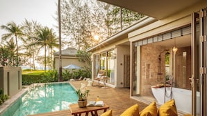 A pool area with a wooden deck, yellow cushions, a table with a book and flowers, and a view of a spa and greenery. - Devasom Khao Lak Beach Resort & Villas (Takua Pa)