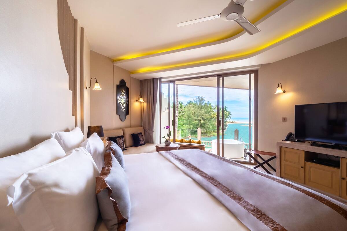 A hotel room with a large bed, a TV, a balcony with a view of the sea, and a ceiling fan.