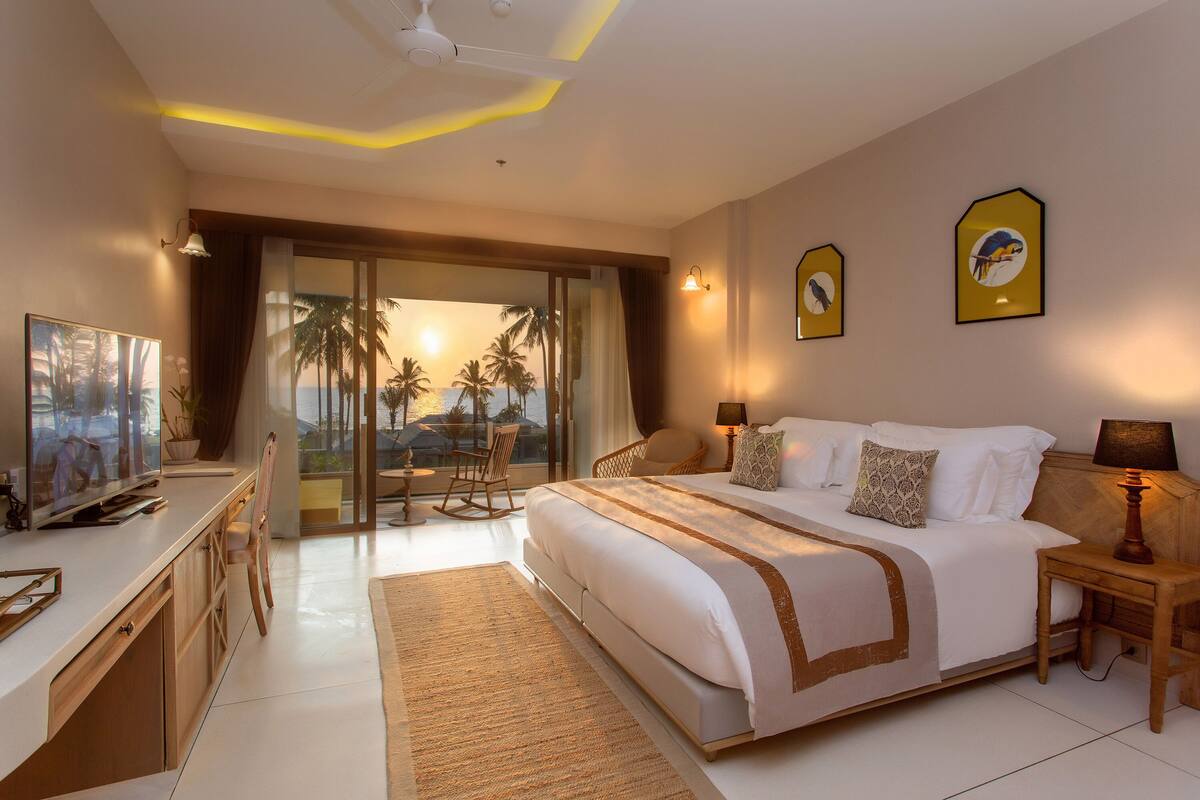A spacious bedroom with a large bed, a desk with a television, and a view of palm trees through a sliding door.