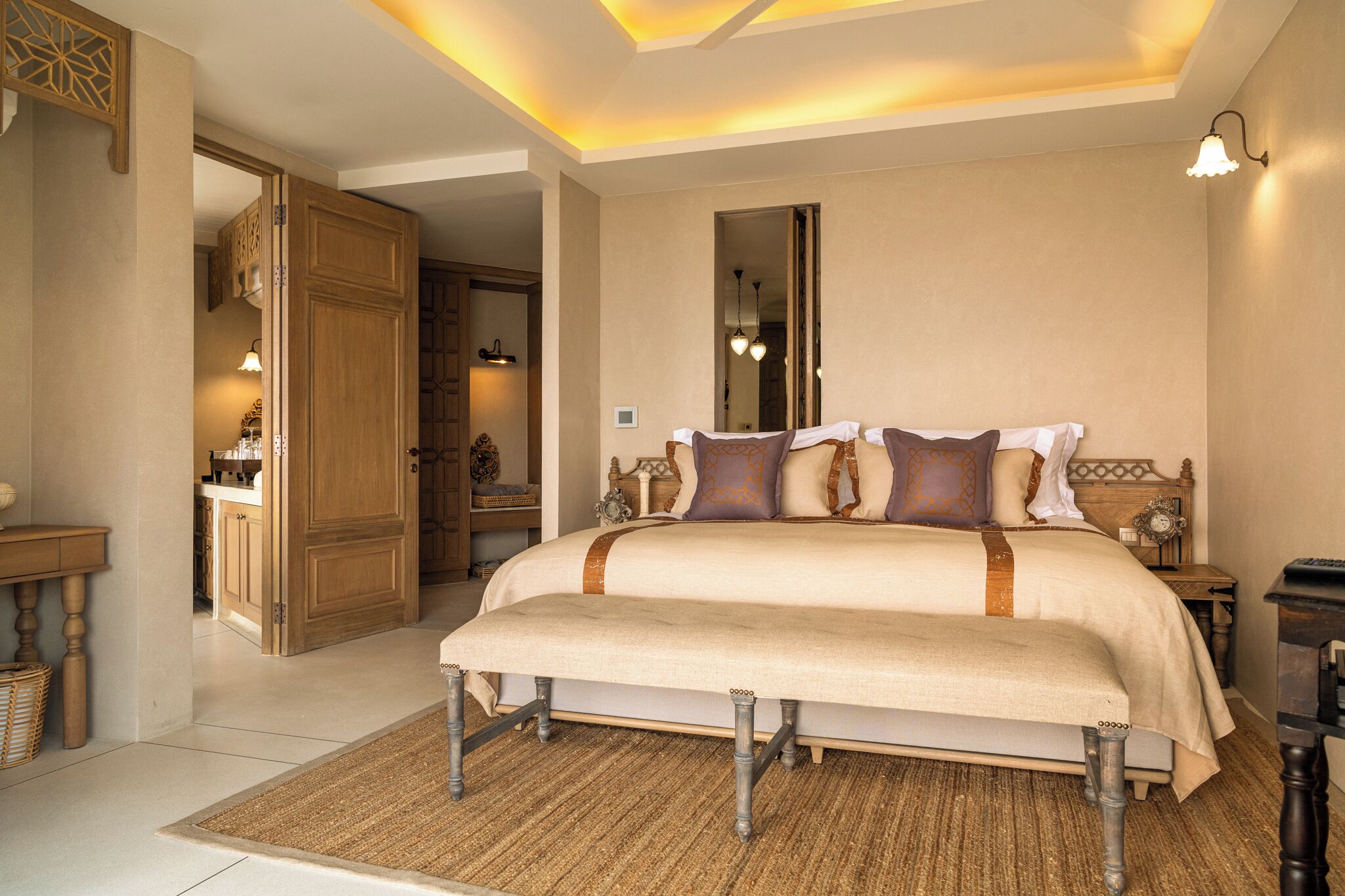 A bedroom with a large bed, wooden headboard, bedside tables, a nightstand, a mirror, and a door leading to a bathroom.