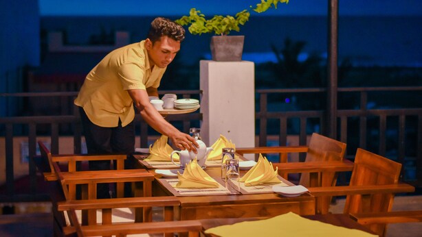 A person setting up a table with yellow napkins and a teapot on a wooden table outdoors at night. - Asian Grand Hotel (Negombo)