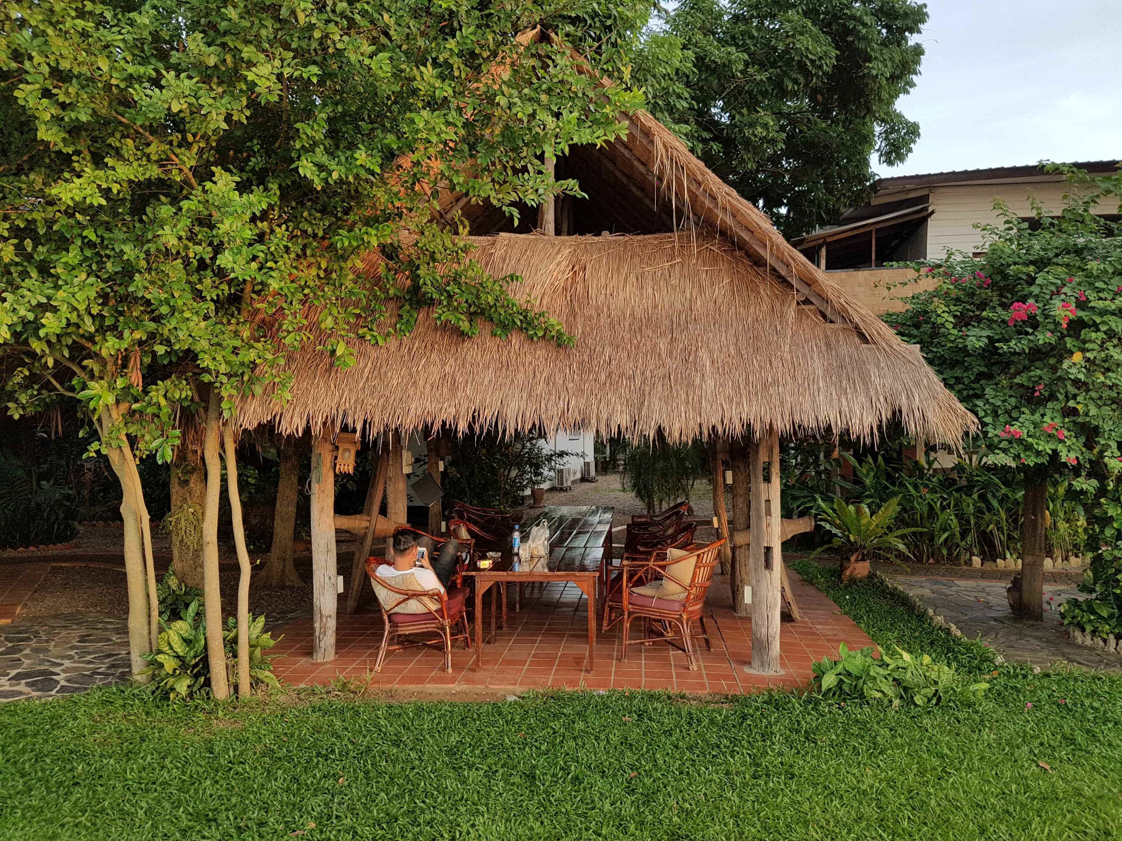 A thatched-roof outdoor seating area with wicker chairs and a table, surrounded by lush greenery and trees.