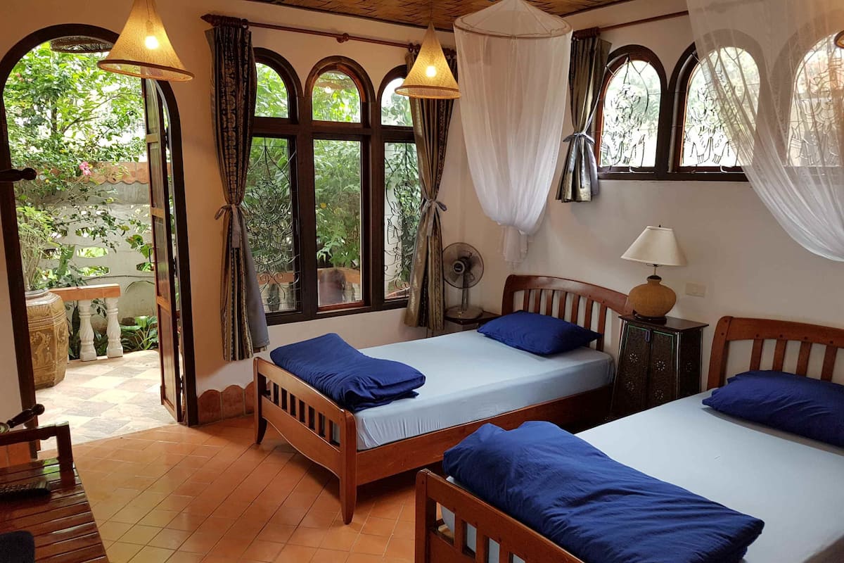 Habitación con dos camas de madera, cada una con ropa de cama azul, una mesita pequeña, una silla y un ventilador.