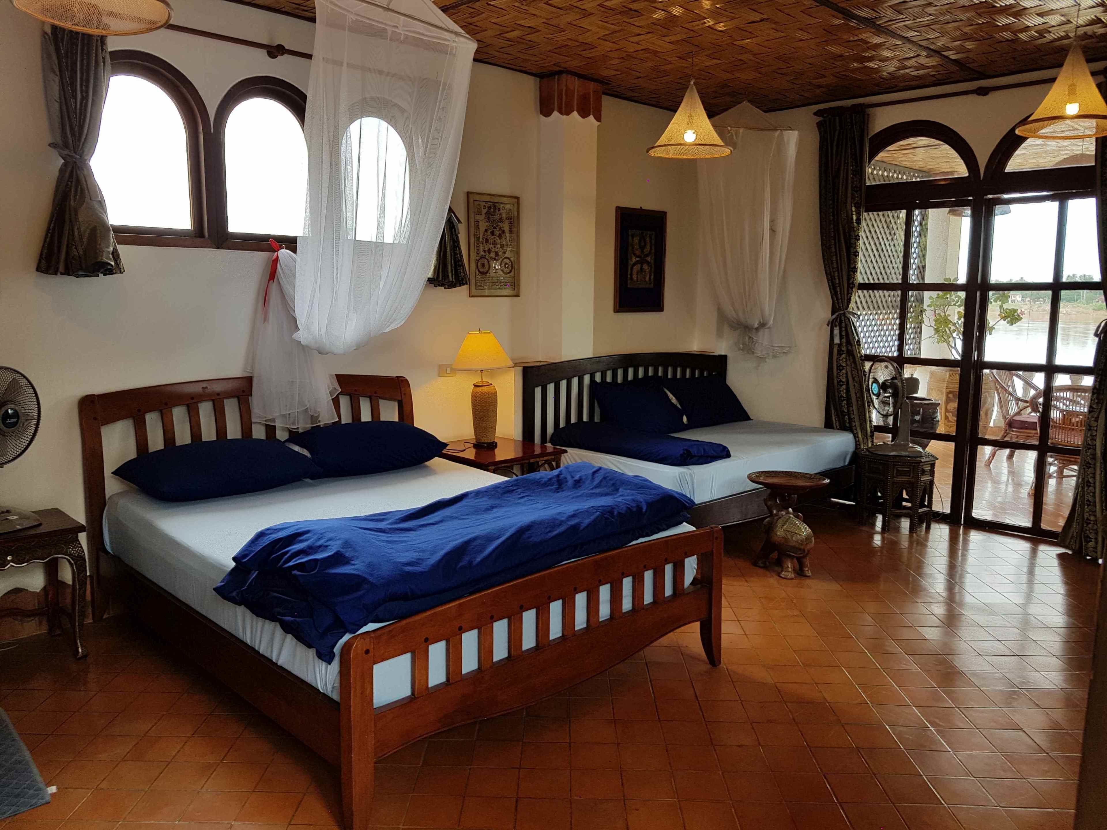 A room with two wooden beds, a small table, a standing fan, and a window with curtains.
