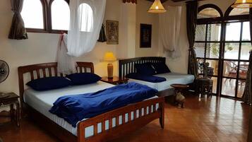 A room with two wooden beds, a small table, a standing fan, and a window with curtains.