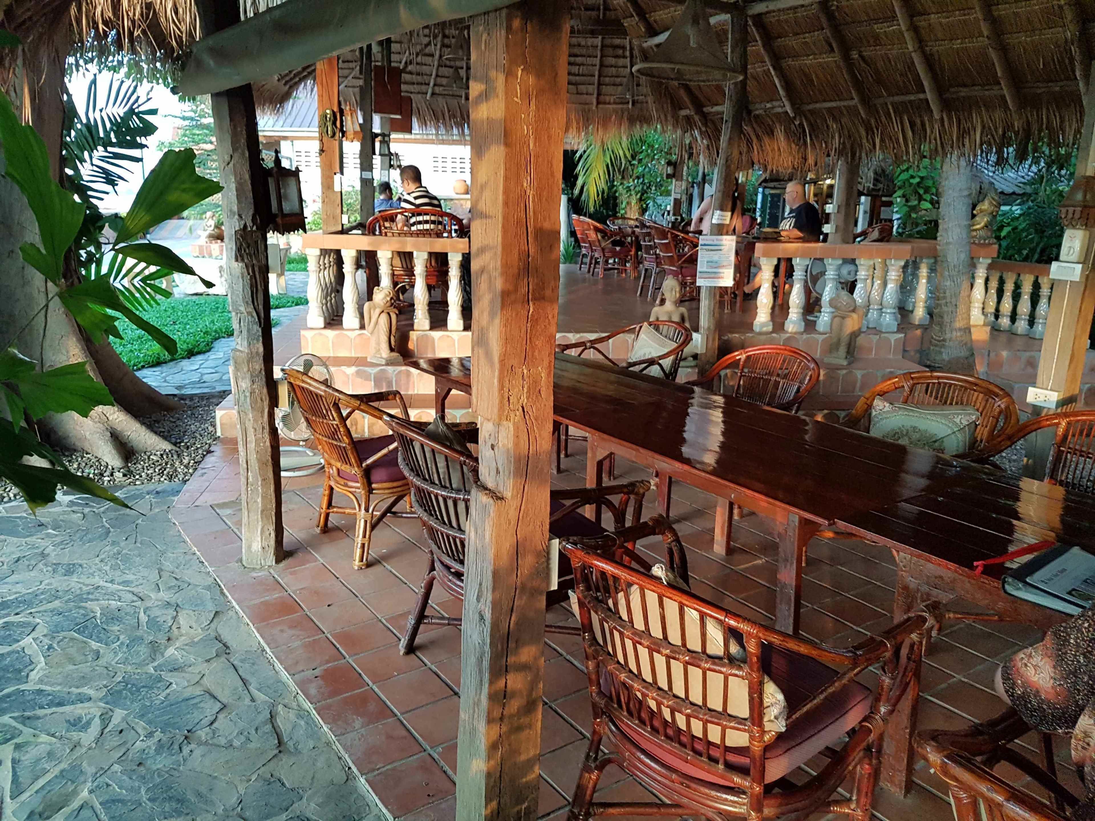 An outdoor dining area with wooden tables and chairs, surrounded by a thatched roof structure.