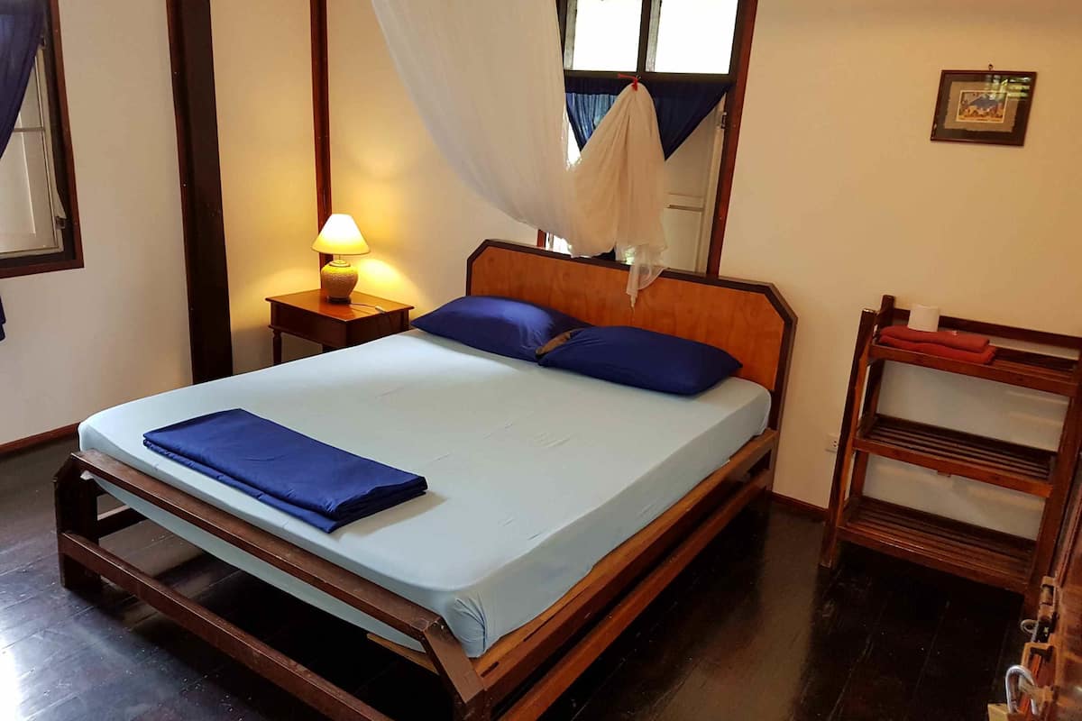 Una cama bien hecha con ropa de cama y almohadas azules, una lámpara de noche, una mesita de madera y un banco de madera con una toalla y una taza.