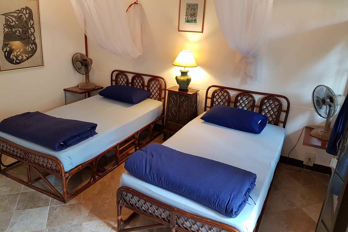 Una habitación con dos camas de madera, cada una con una almohada azul y una colcha blanca. Hay dos mesitas de noche con lámparas y ventiladores.