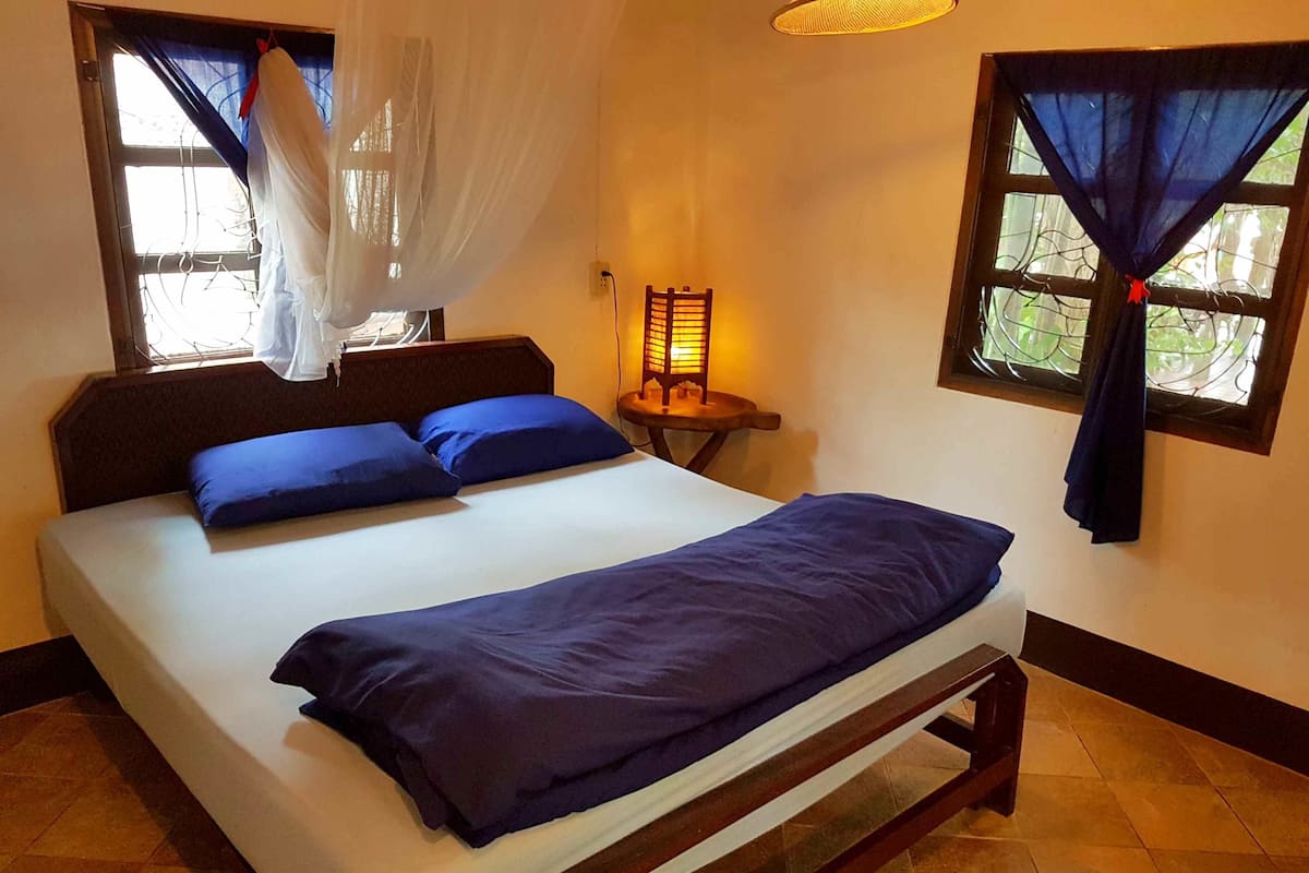 Una cama con ropa de cama azul, una mesita de noche con una lámpara y ventanas con cortinas.