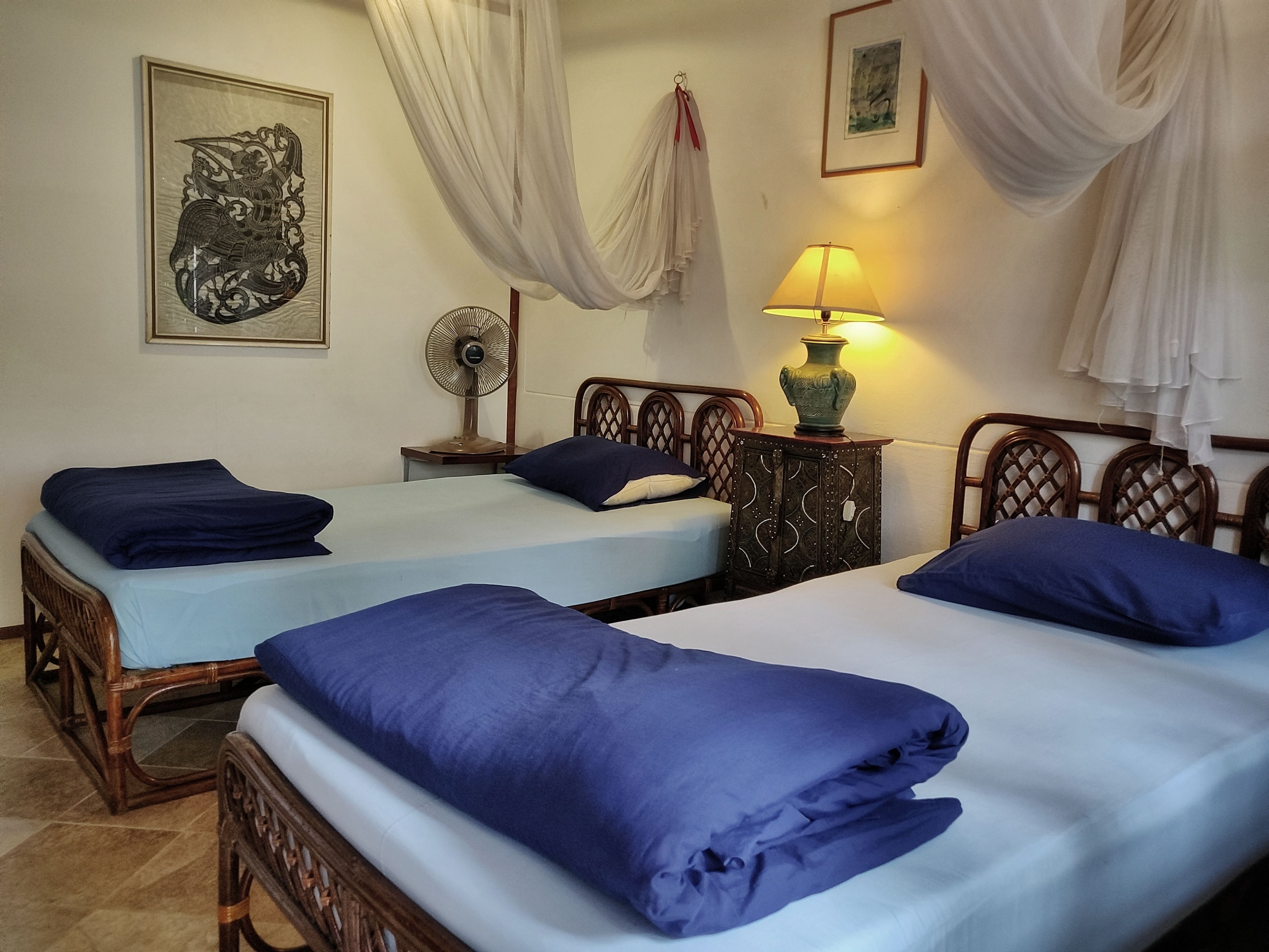 Habitación de hotel con dos camas, una mesita de noche con lámpara, un ventilador y un adorno en la pared.