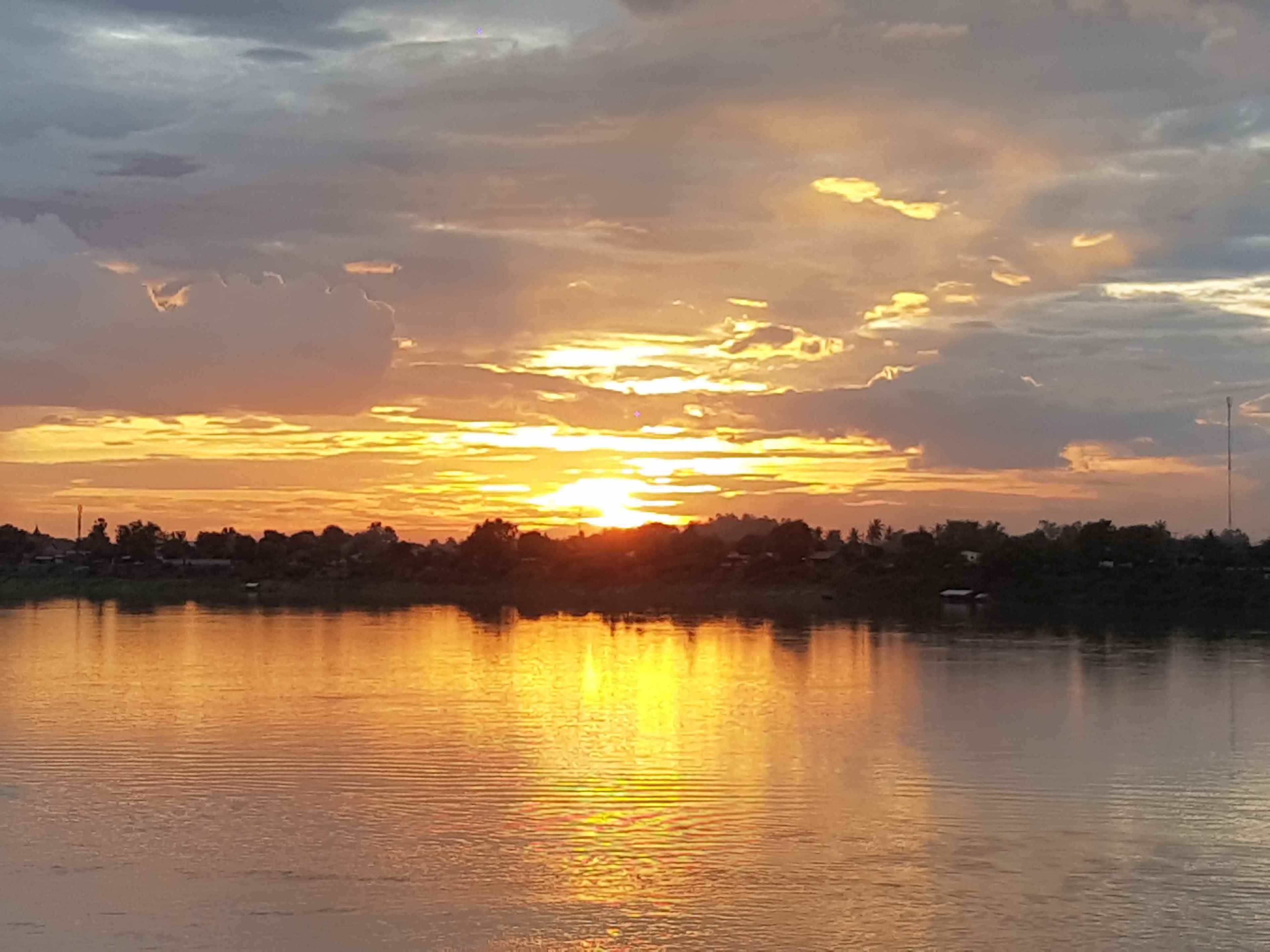 Un coucher de soleil sur un lac paisible, avec en arrière-plan la silhouette d’arbres et de bâtiments.