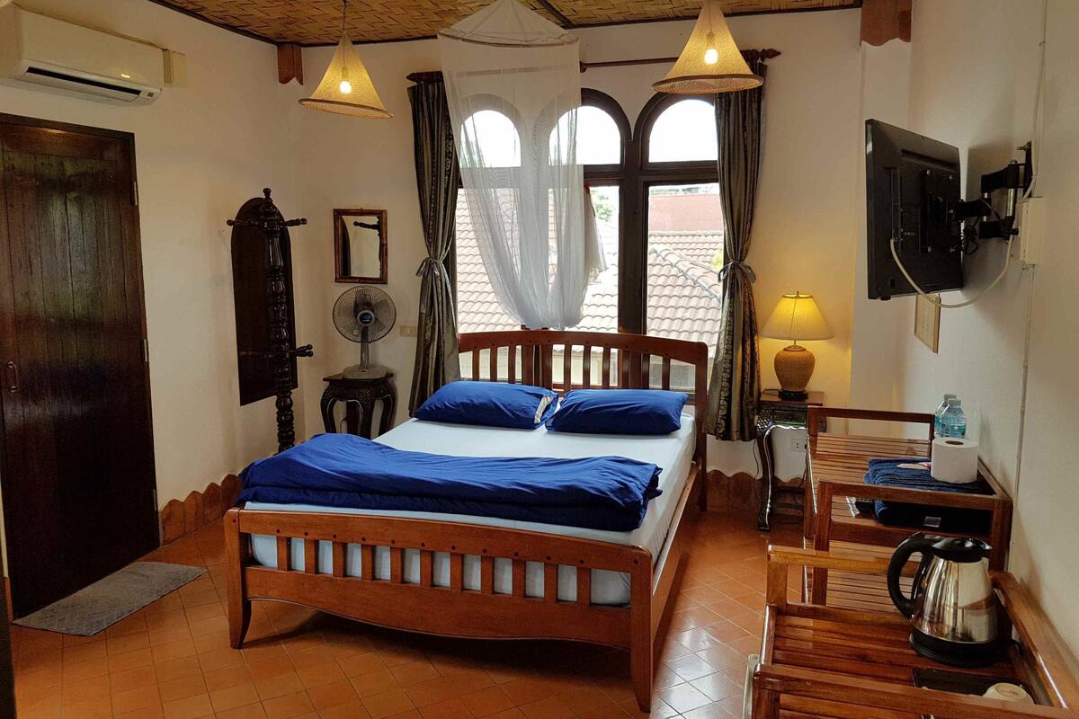 Un dormitorio con una cama de madera, una mesita de noche con lámpara, un televisor montado en la pared y un ventanal con cortinas.